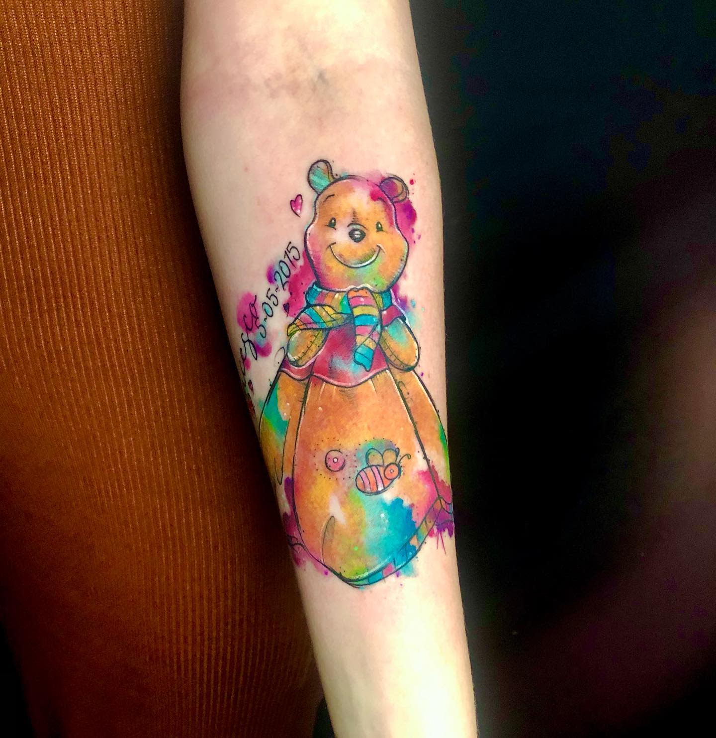 Colorful tattoo of Winnie the Pooh wearing a scarf with a butterfly and bee on forearm